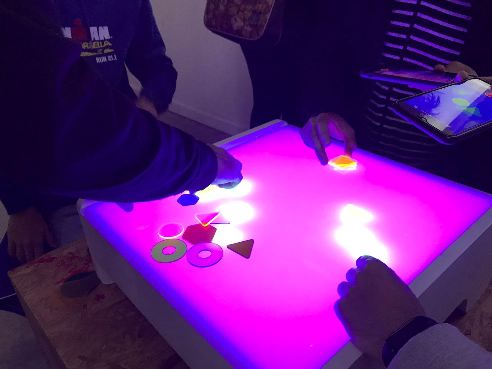 En primer plano una mesa con luz fluorescente iluminandola desde abajo y tres niños realizando actividades con piezas geometricas de plastico