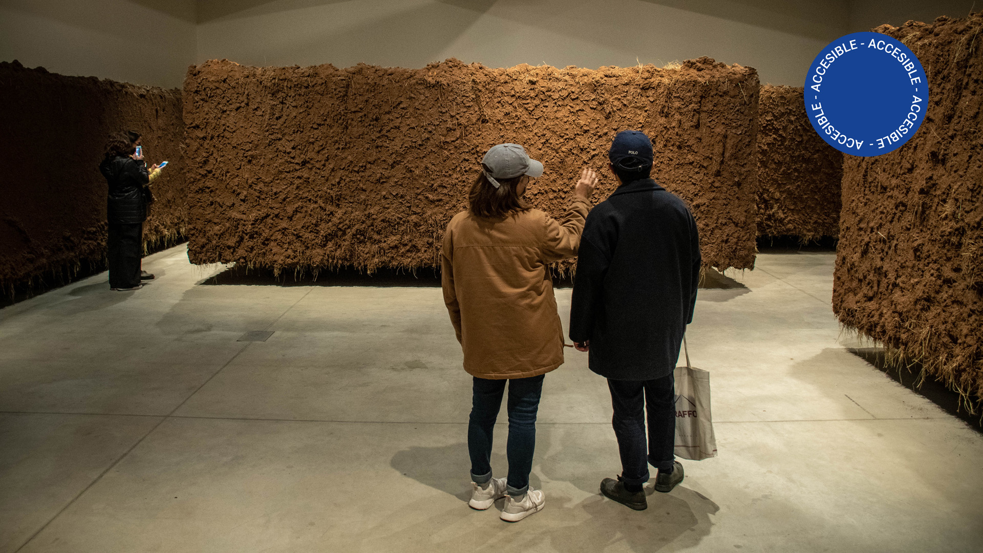 Imagen fotográfica que muestra a personas en la sala de la exposición observando las paredes de tierra que conforman la obra y sacando fotos desde un celular.