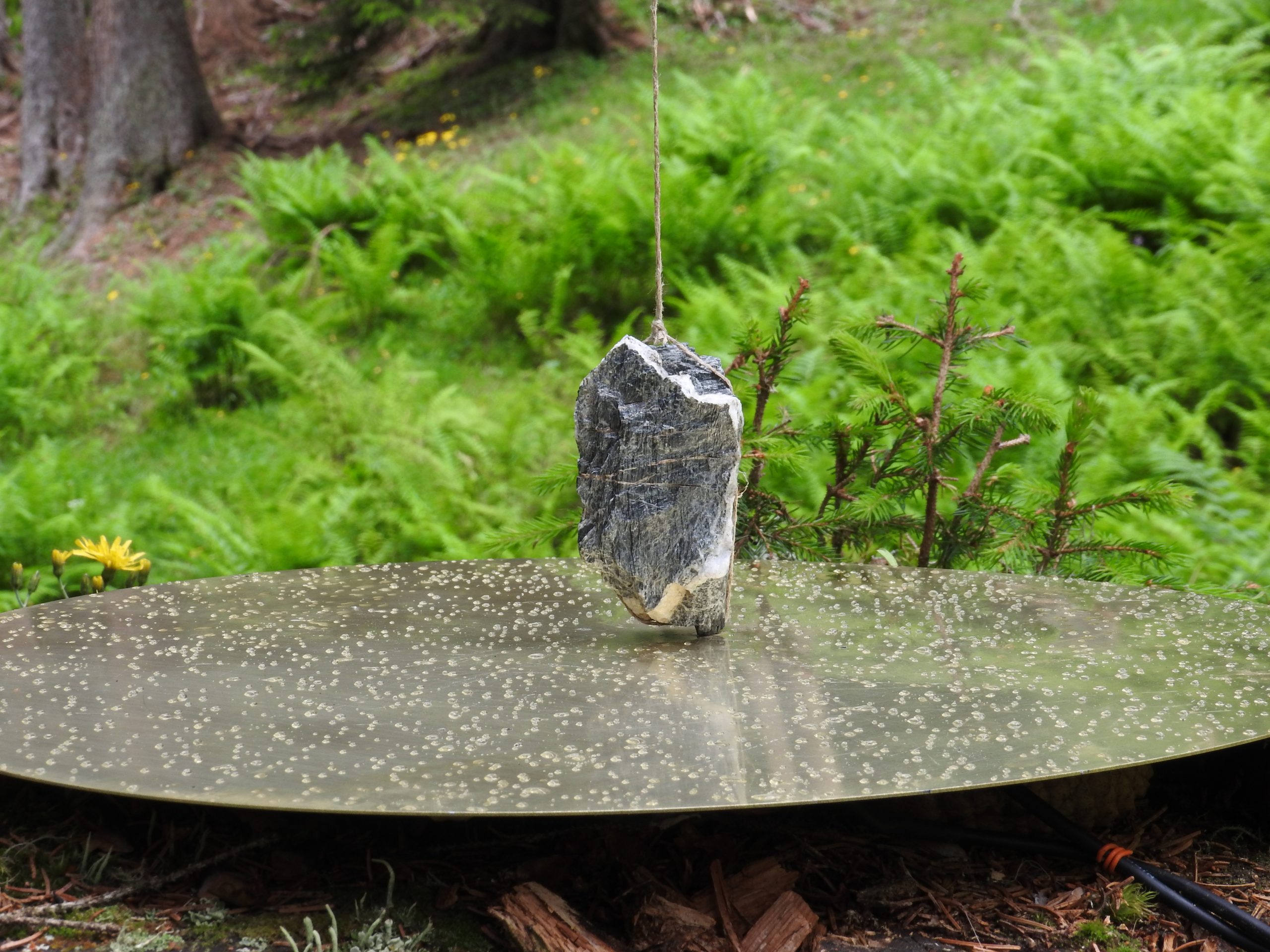 La fotografía muestra una piedra atada de un hilo, apoyada como péndulo sobre un disco plano agujereado, con un paisaje frondoso de fondo.
