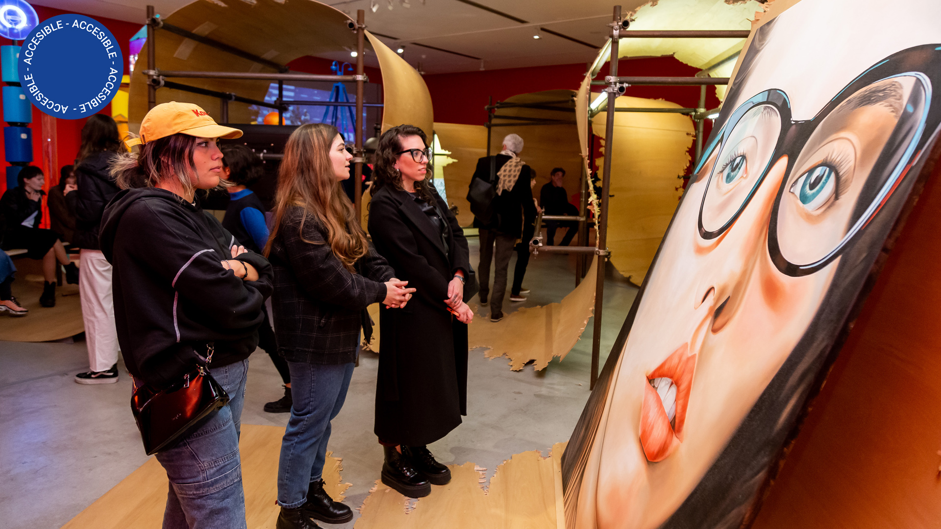 La imagen muestra una pintura en primer plano –el retrato de una mujer con gafas– y a tres mujeres comentando enfrente de la obra. De fondo, se ven personas recorriendo la exposición.