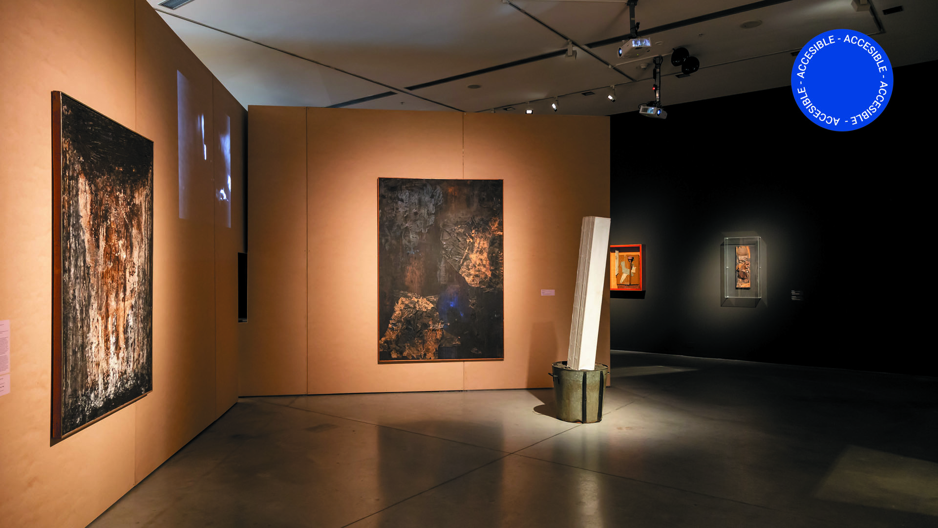 La imagen muestra una foto de la sala en donde se ven grandes cuadros colgados sobre paneles en tonos oscuros y con iluminación tenue.