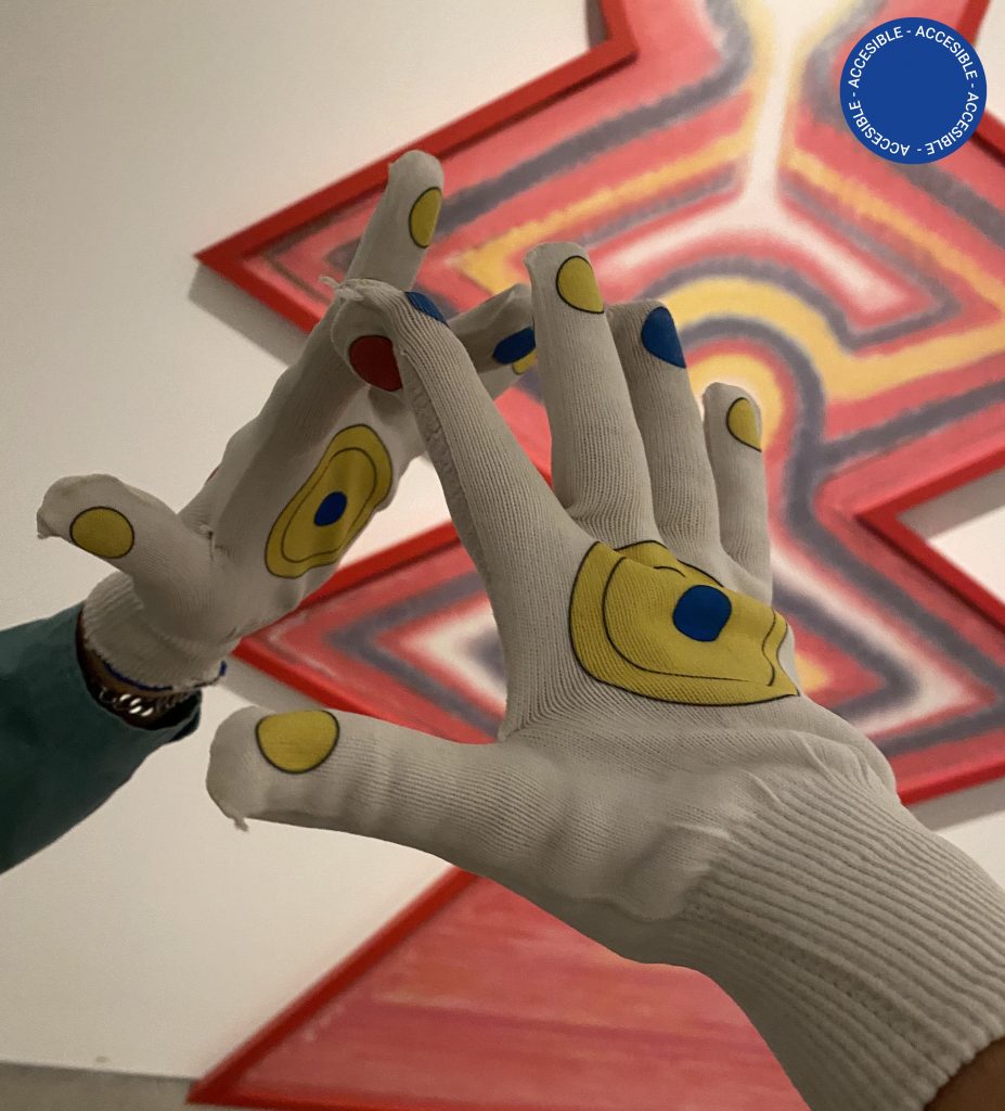 Imagen dos manos vistiendo el dispositivo sensorial usado en la obra Torres magnéticas, de Eduardo Navarro. Se ven dos guantes en primer plano, la obra sobre la pared de fondo.