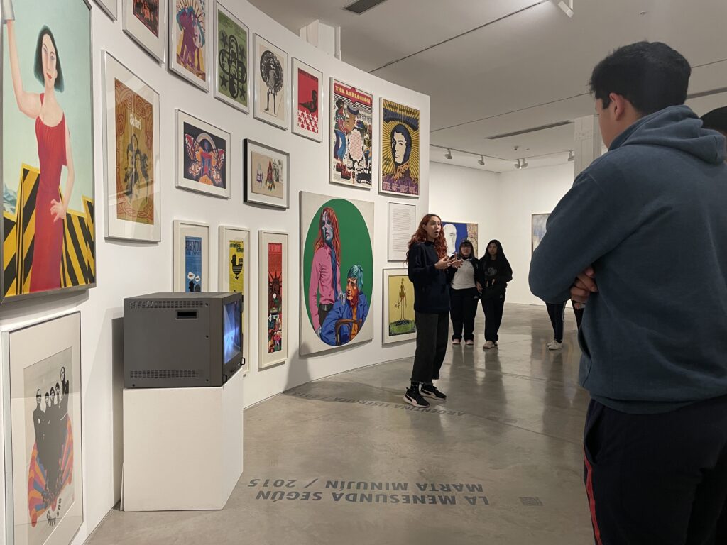 Fotografía tomada en la sala de exposición donde se ve la educadora del Museo frente a un grupo de visitantes. De fondo, hay pinturas coloridas de diferentes tamaños sobre una pared curva.