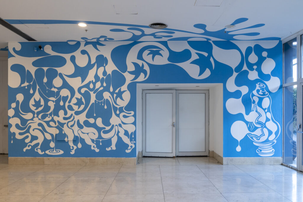 La imagen muestra el hall principal del Moderno, donde se encuentra la puerta de acceso a la sala Yapeyú, intervenido con un mural monocromático en color turquesa. Se trata de la exposición Alberto Passolini: Soñar a borbotones, en la cual, a partir de una gota, se despliega un mural que ocupa todas las paredes y el techo del lugar.