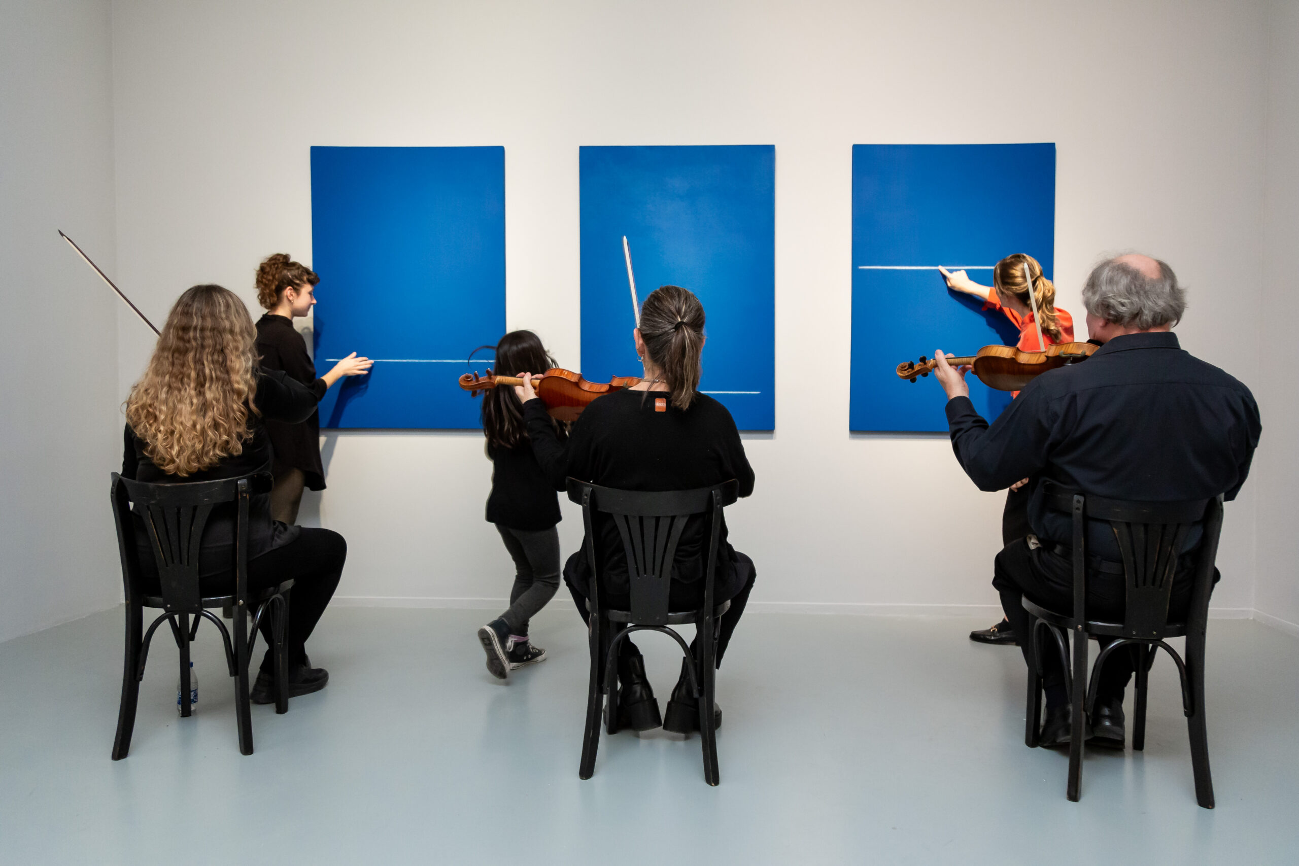 La fotografía muestra una activación en sala, en la que tres personas siguen con sus manos el recorrido de las líneas blancas que atraviesan tres cuadros de color azul. Detrás, tres músicos interpretan sonidos con sus violines.