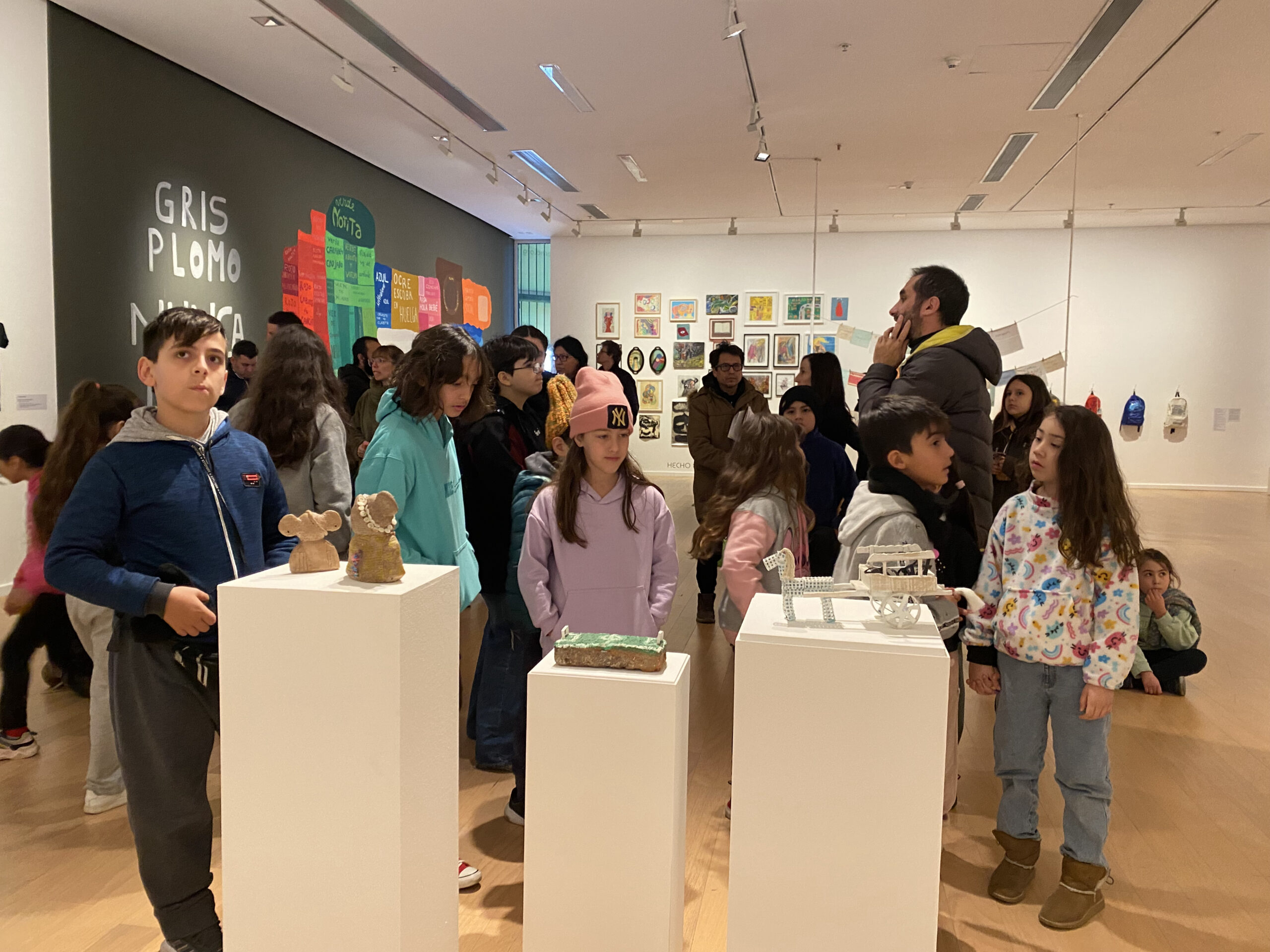 La imagen muestra una visita a la sala de exposición del Museo. En primer plano, se ve a un grupo de niños que observan objetos de arte exhibidos en pequeñas tarimas. En el fondo, hay otras obras en sala y públicos diversos.