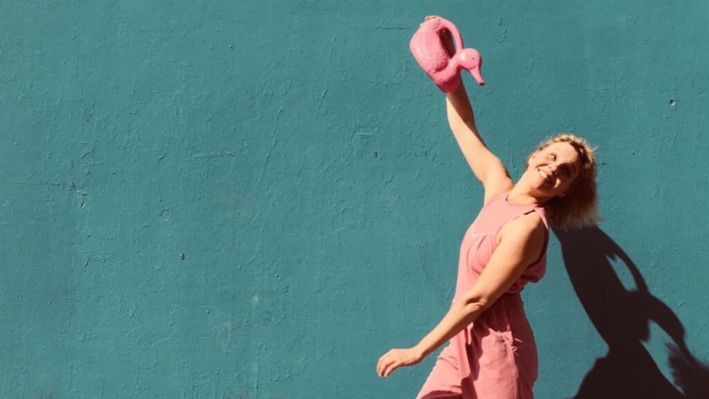 Mujer sonriente en overol rosa, jugando con una regadera sobre una bañera colorida