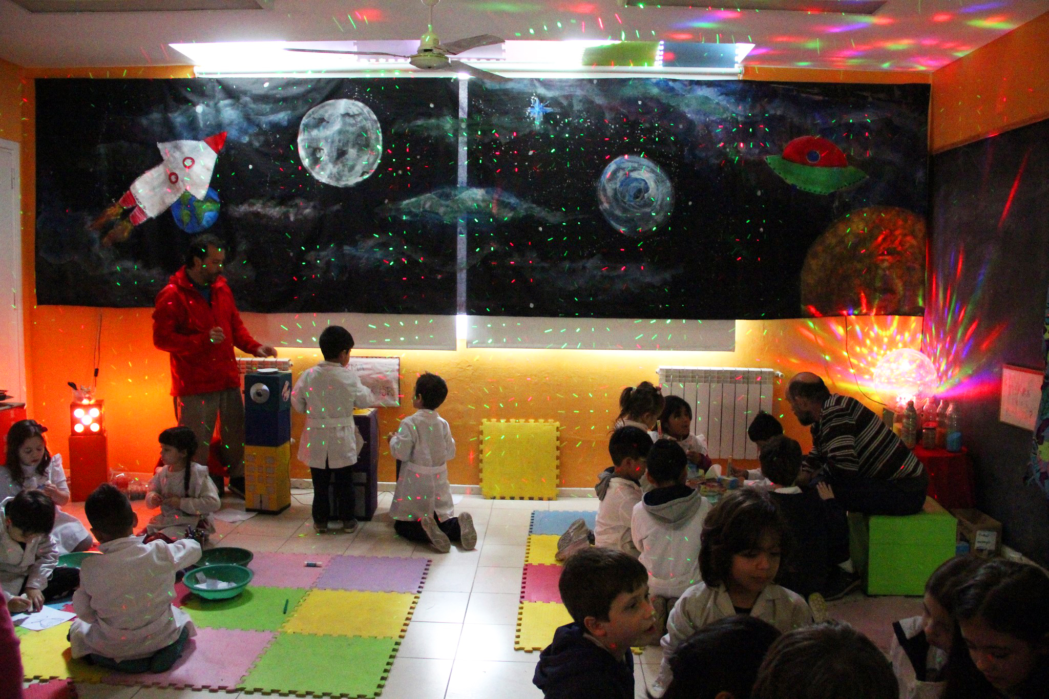 En la imagen se observa un grupo de niños sentados en un taller con distintas alusiones al espacio exterior