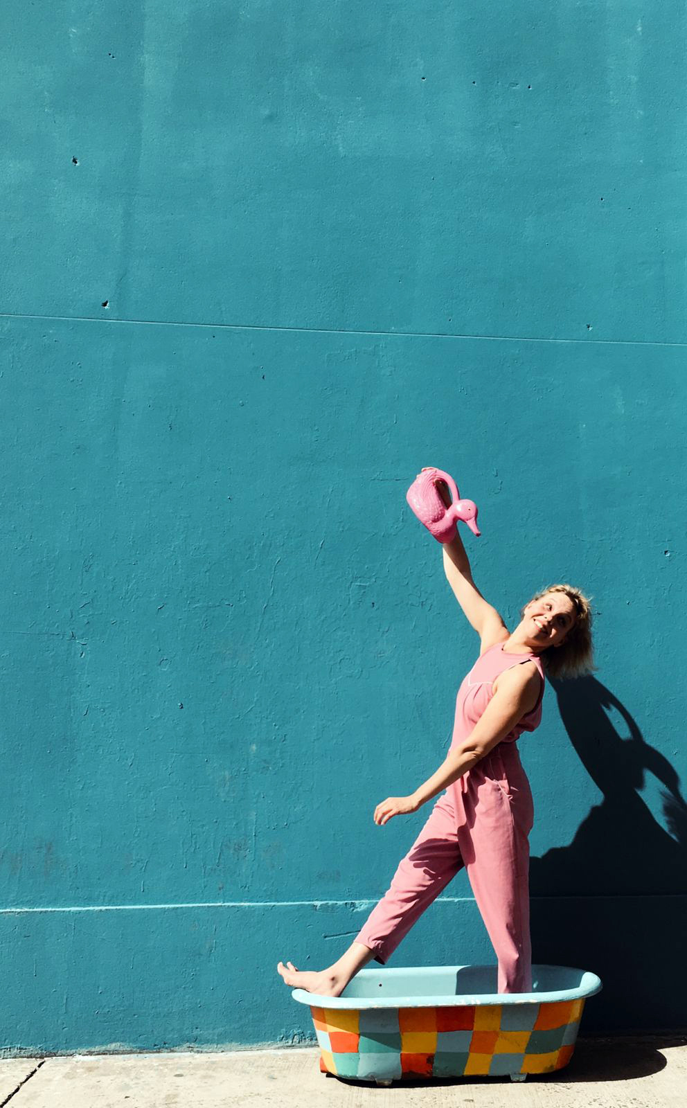 Una mujer sonriente, en overol rosa, juega con una regadera sobre una bañera colorida.