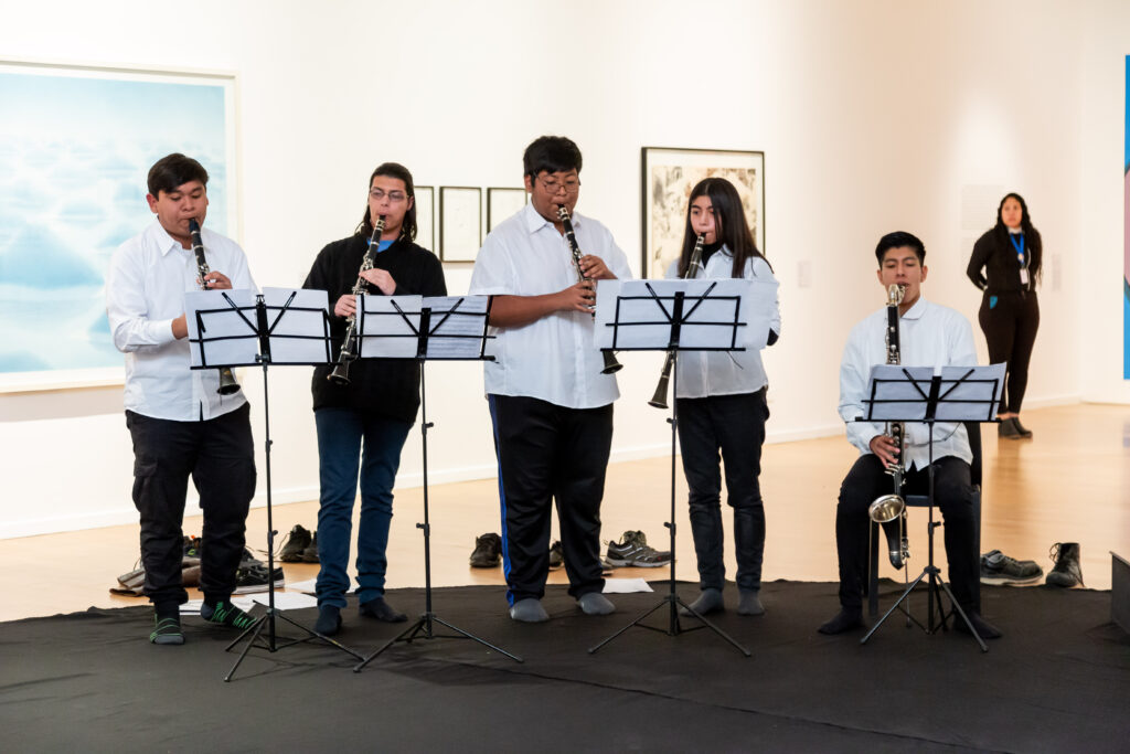 La imagen muestra un grupo de jóvenes tocando instrumentos en una sala de exposiciones del Museo.