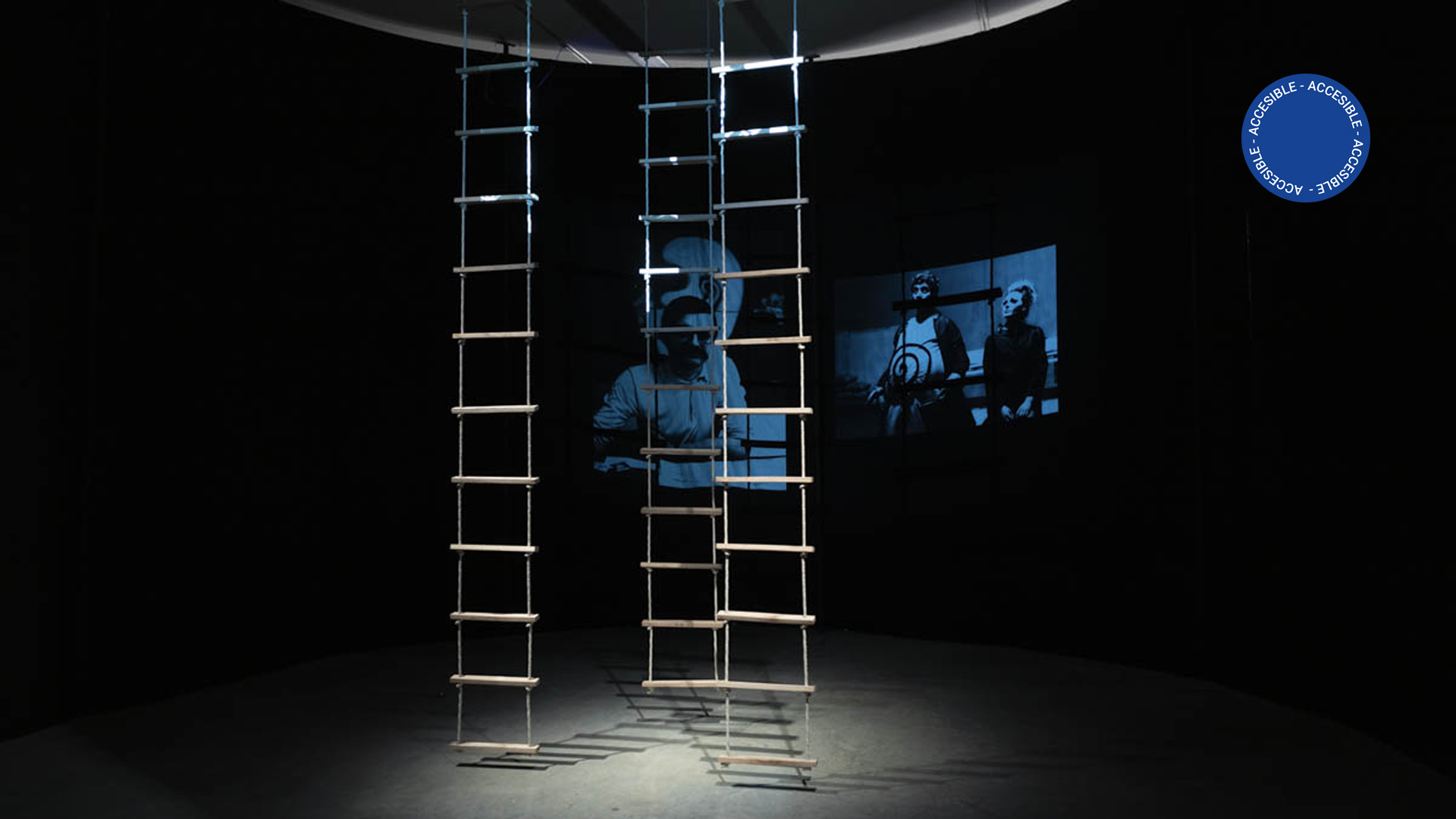 La imagen muestra un espacio circular delimitado por una gran cortina negra sobre la cual se proyectan imágenes en tonos azulados. En el centro del espacio hay tres escaleras de nudos, como las que pueden encontrarse en antiguas embarcaciones.