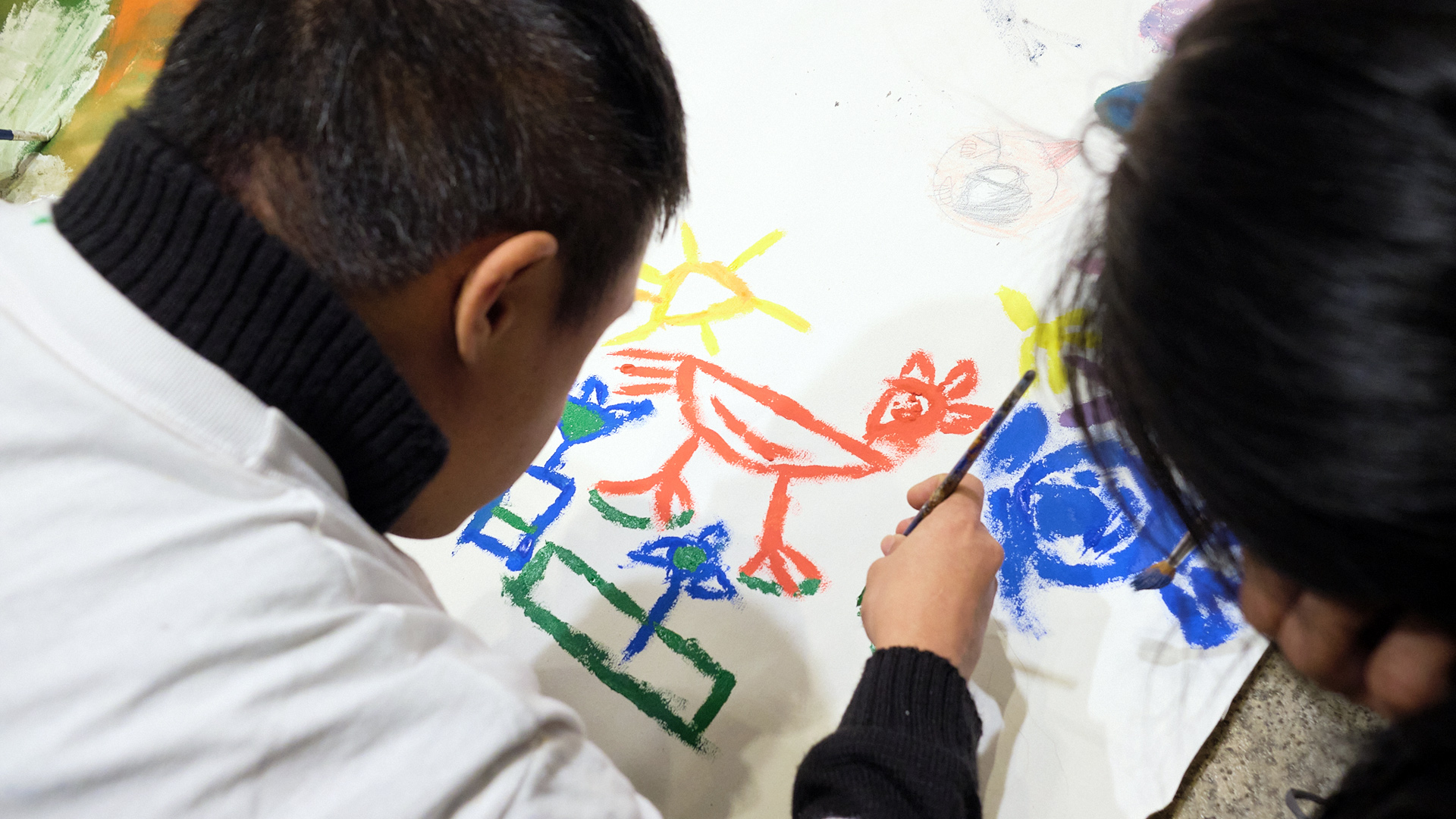 La imagen muestra en primer plano la pintura que está realizando una persona que se ve de espaldas, otra está a su lado.