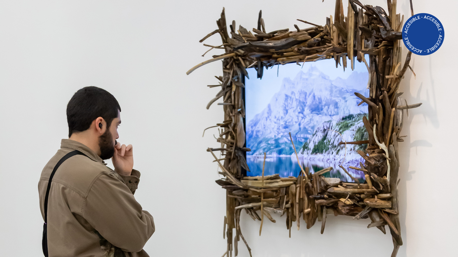 La foto muestra a una personas de espaldas viendo una TV de pantalla plana donde se proyecta un paisaje. El marco de la TV está cubierto de pequeños tronquitos de madera. La pieza es parte de la instalación de Antonio Villa en la sala de exposición del segundo subsuelo del Moderno.