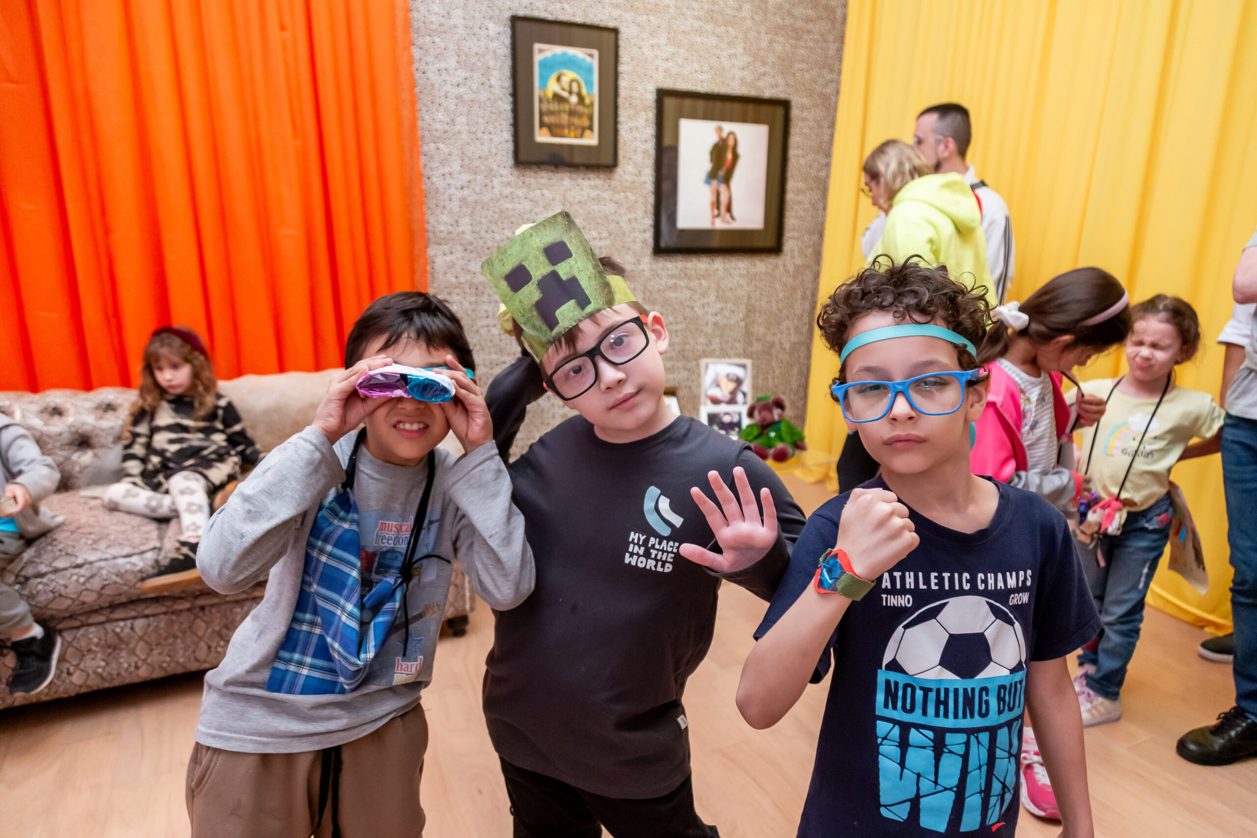 En la fotografía a color se observa un grupo de niños y tres de ellos de frente a la cámara, mostrando sombreros, pulseras y lentes realizados en uno de los varios talleres artístico-educativos que gratuita y públicamente el Museo de Arte Moderno de Buenos Aires realiza periodicamente, dentro de la exposición Dalila Puzzovio: Autorretrato.