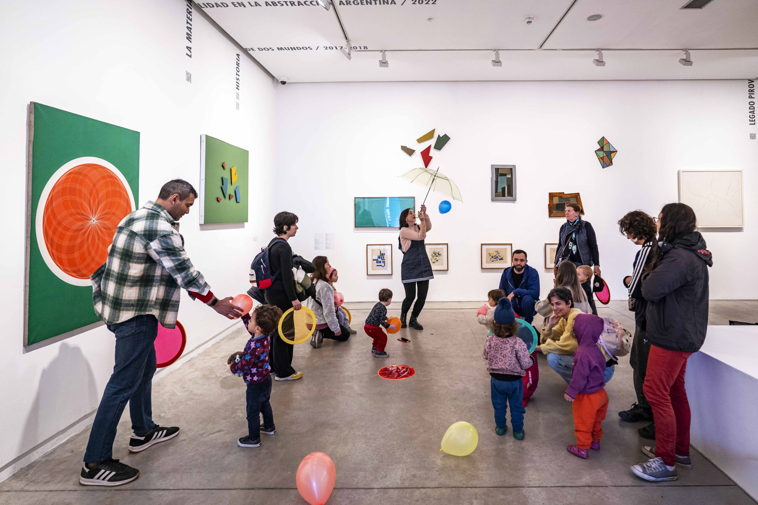 En la fotografía a color se observan varios grupos familiares compuestos por madres, padres y niños que con la guía de un educador estan realizando una serie de talleres y juegos con globos de colores y otro tipo de apoyos dentro de la exposición Moderno y MetaModerno.