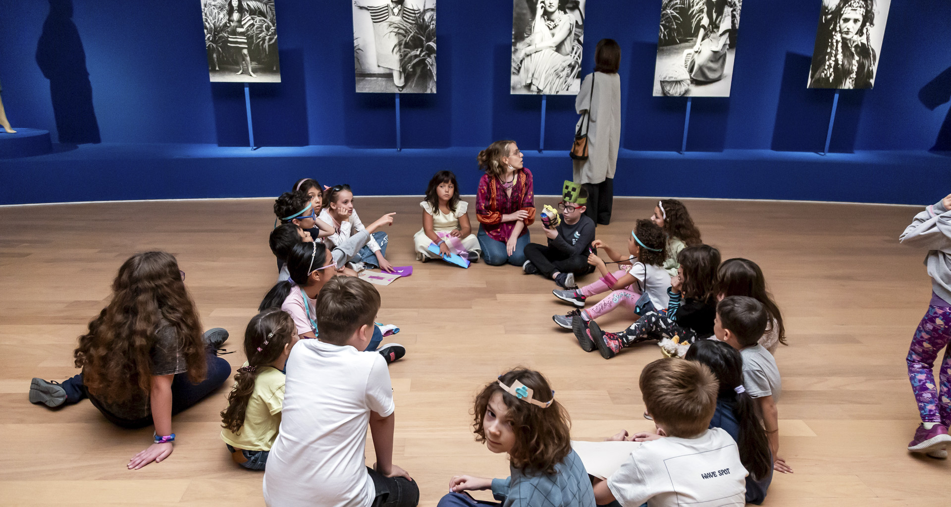 En la fotografía a color un grupo de niños se sientan en ronda sobre el piso de la exposición Dalila Puzzovio: Autorretrato en la que destacada una de las educadoras del Museo Moderno en la que está señalando una obra sobra la cual debe estar enseñandole a los infantes.