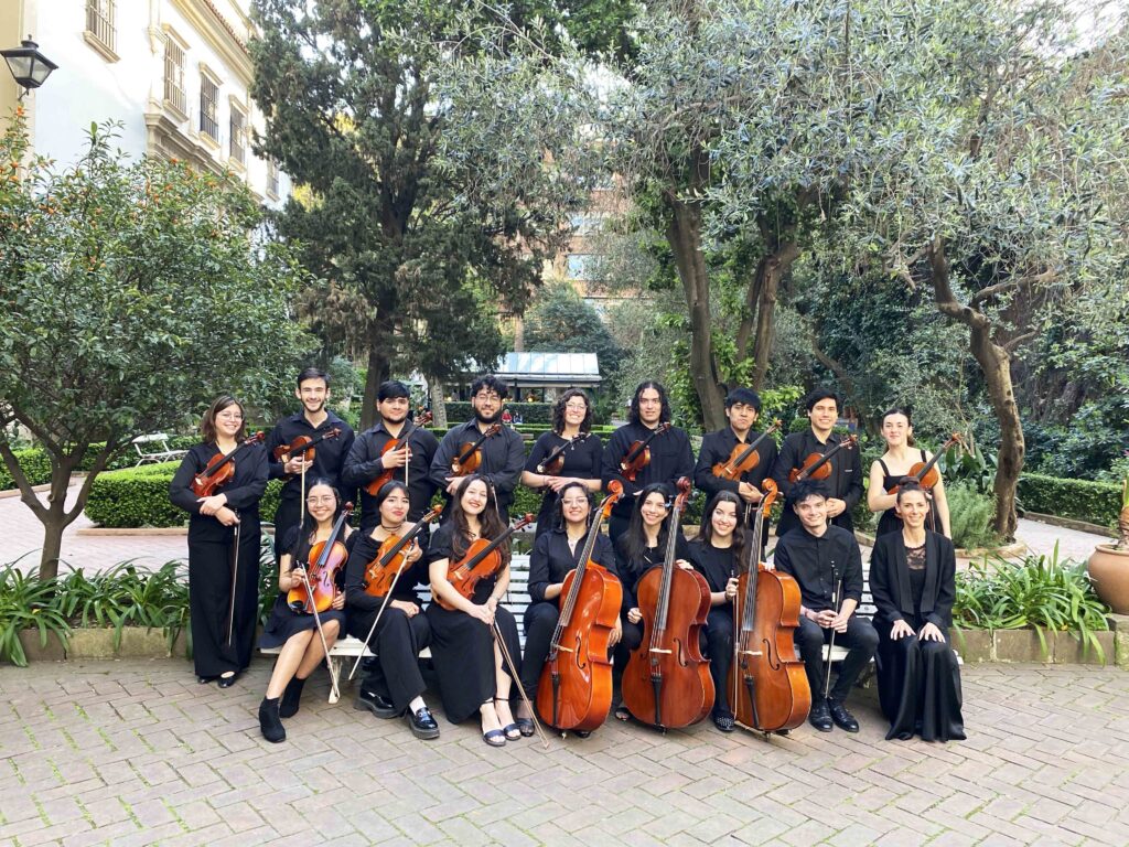 La imagen muestra al grupo integrado por 16 jóvenes músicos vestidos de negro que posan, formados, junto a su directora. Están en un patio con muchas plantas y árboles, donde los instrumentos musicales se destacan por su brillo y su gran tamaño en el caso de los contrabajos.