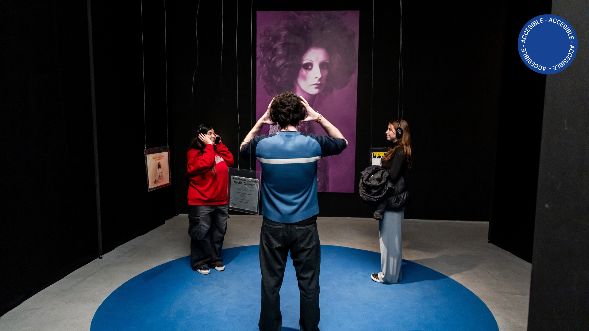 En la fotografía a color se puede observar a tres jóvenes escuchando con auriculares una obra de la exposición Esto es teatro. Once escenas experimentales: del Di Tella al Parakultural