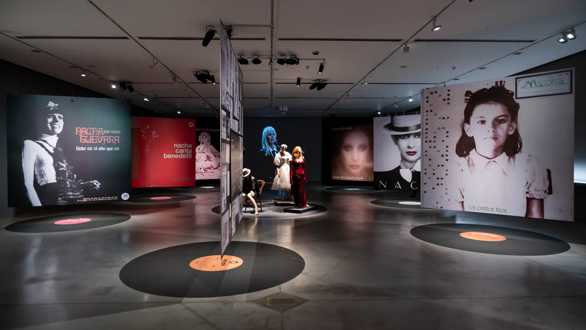 La imagen muestra la sala de la exposición Nacha Guevara: Aquí estoy donde se ven grandes paneles con fotografías de la artista, suspendidas arriba de unos discos de vinilo pegados en el suelo. En el centro de la imagen, se ven maniquíes con ropas, y de fondo, un video de gran formato donde se ve a Nacha cantando.