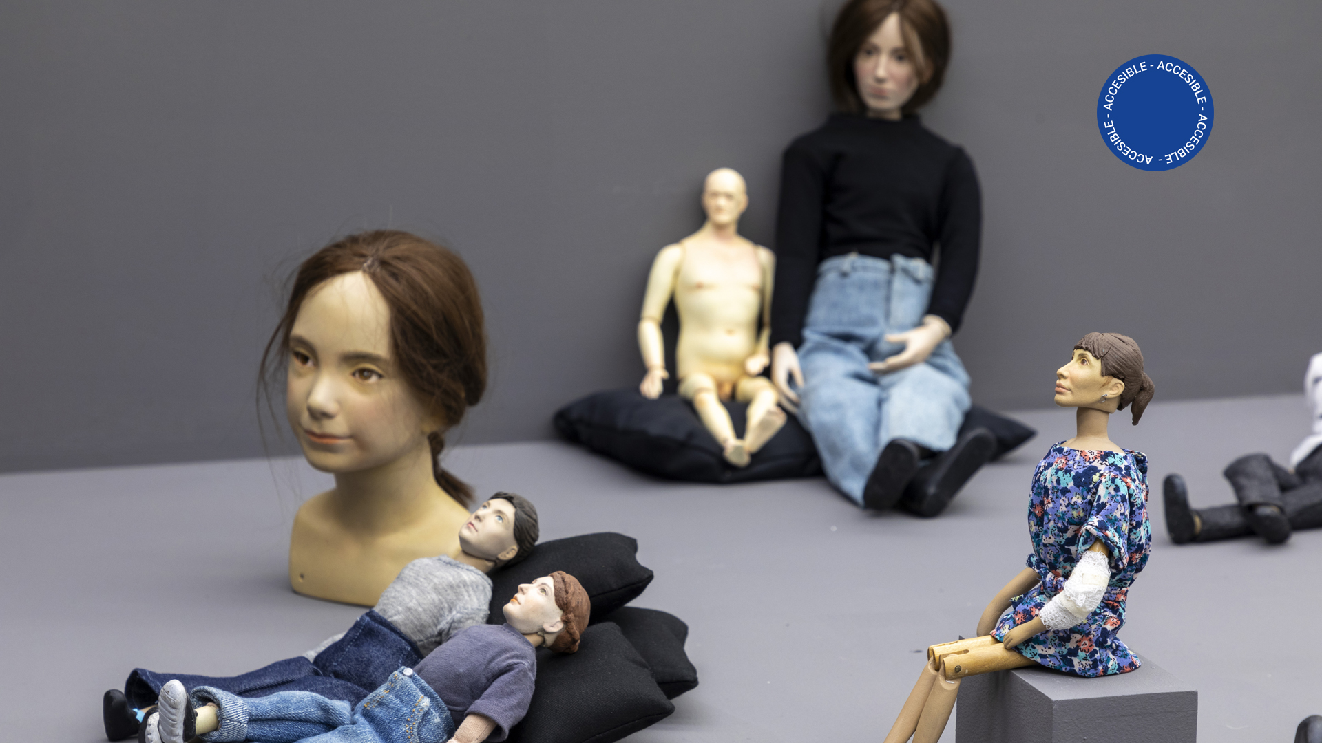 La imagen muestra muñecas de porcelana de diferentes tamaños; algunos de estos personajes tienen ropas y otros no. Están sentados o recostados sobre una base gris o sobre pequeños almohadones negros.