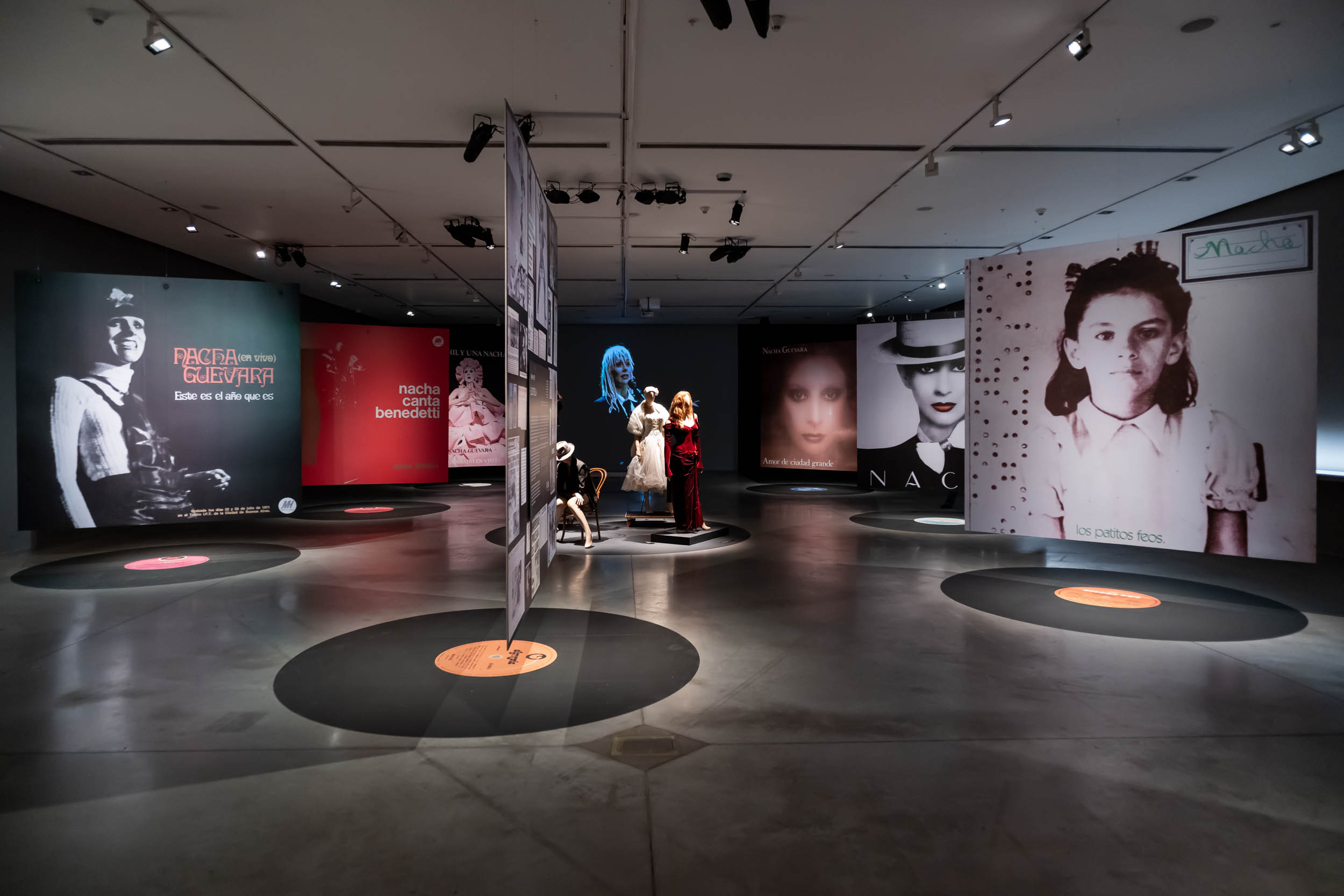 La imagen muestra la sala de la exposición Nacha Guevara: Aquí estoy donde se ven grandes paneles con fotografías de la artista, suspendidas arriba de unos discos de vinilo pegados en el suelo. En el centro de la imagen, se ven maniquíes con ropas, y de fondo, un video de gran formato donde se ve a Nacha cantando.