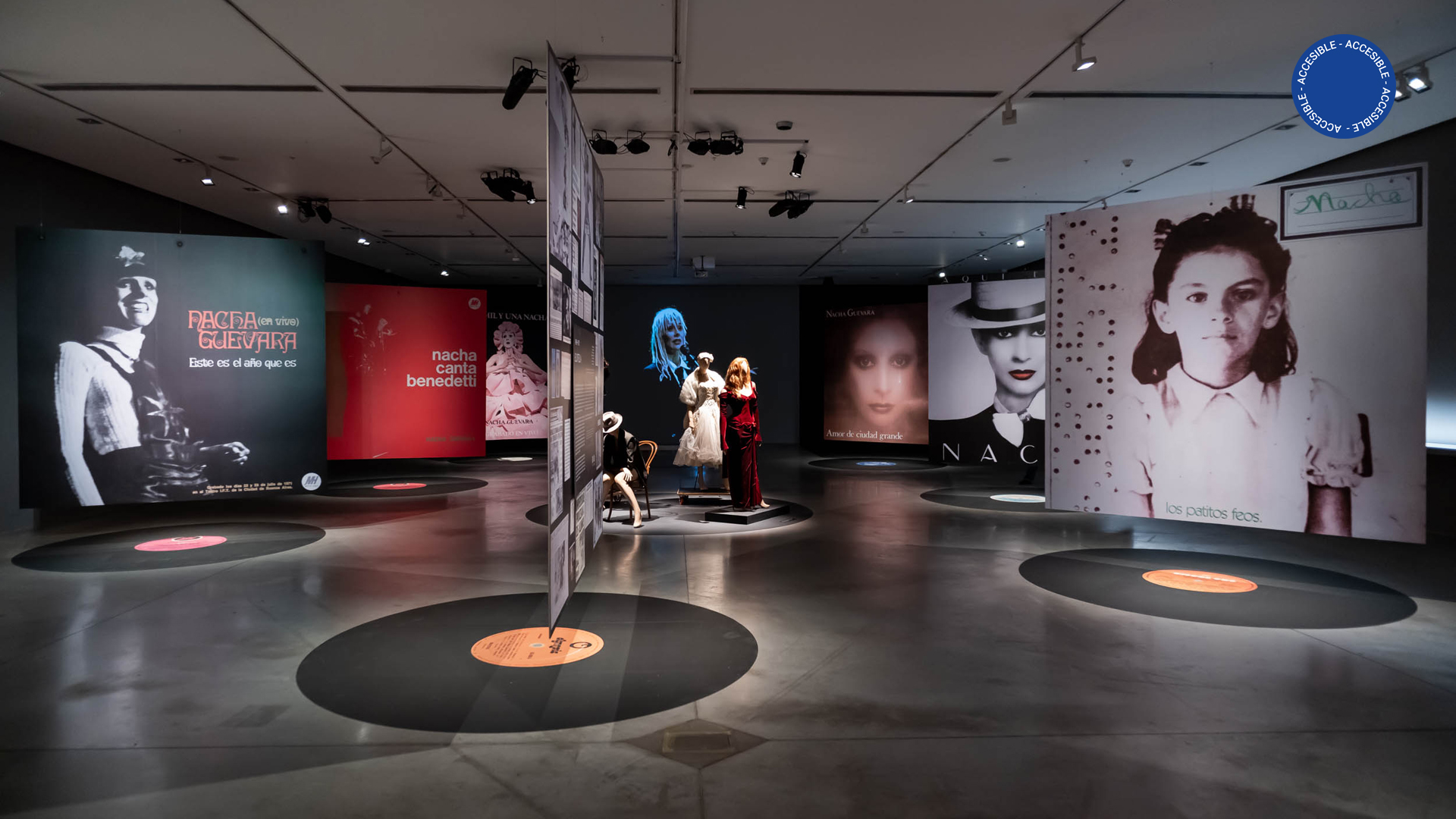 La imagen muestra la sala de la exposición Nacha Guevara: Aquí estoy donde se ven grandes paneles con fotografías de la artista, suspendidas arriba de unos discos de vinilo pegados en el suelo. En el centro de la imagen, se ven maniquíes con ropas, y de fondo, un video de gran formato donde se ve a Nacha cantando.