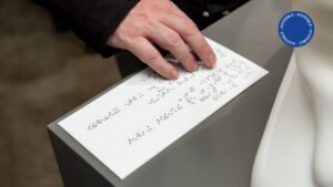 Foto muestra una vista accesible por la sala de educación en la que se ve una mano recorriendo la cédula en Braille de una obra de patrimonio