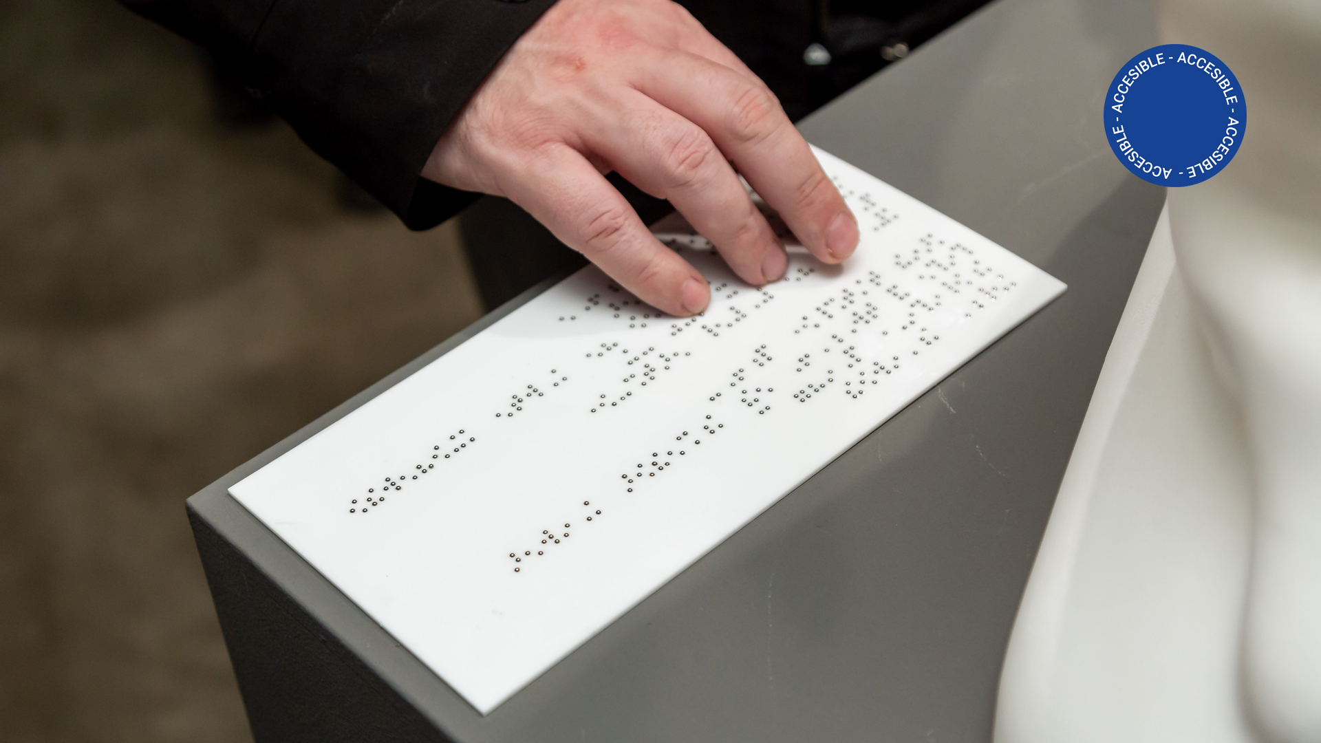 Foto muestra una vista accesible por la sala de educación en la que se ve una mano recorriendo la cédula en Braille de una obra de patrimonio