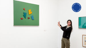 Fotografía a color muestra una sala de la exposición Moderno y MetaModerno donde se exhiben obras de la colección del museo y una persona usando Lengua de Señas.