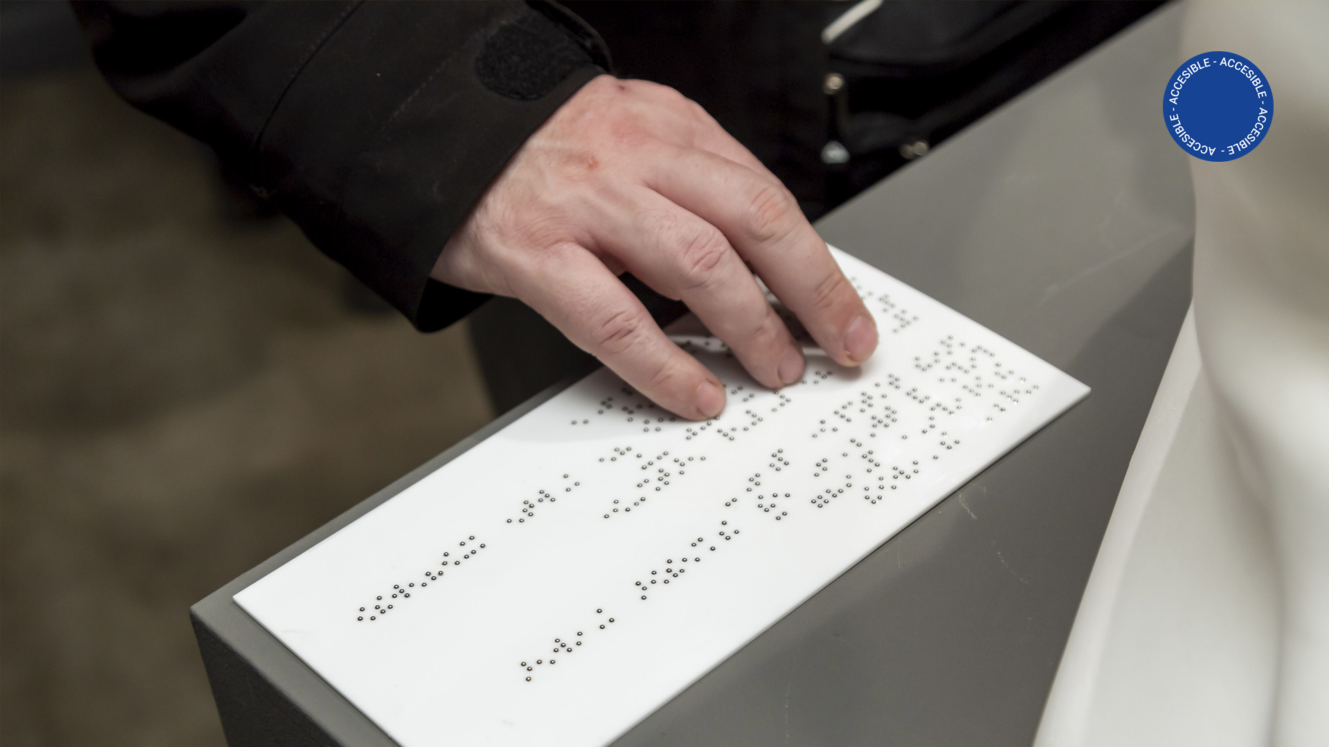 Foto muestra una vista accesible por la sala de educación en la que se ve una mano recorriendo la cédula en Braille de una obra de patrimonio