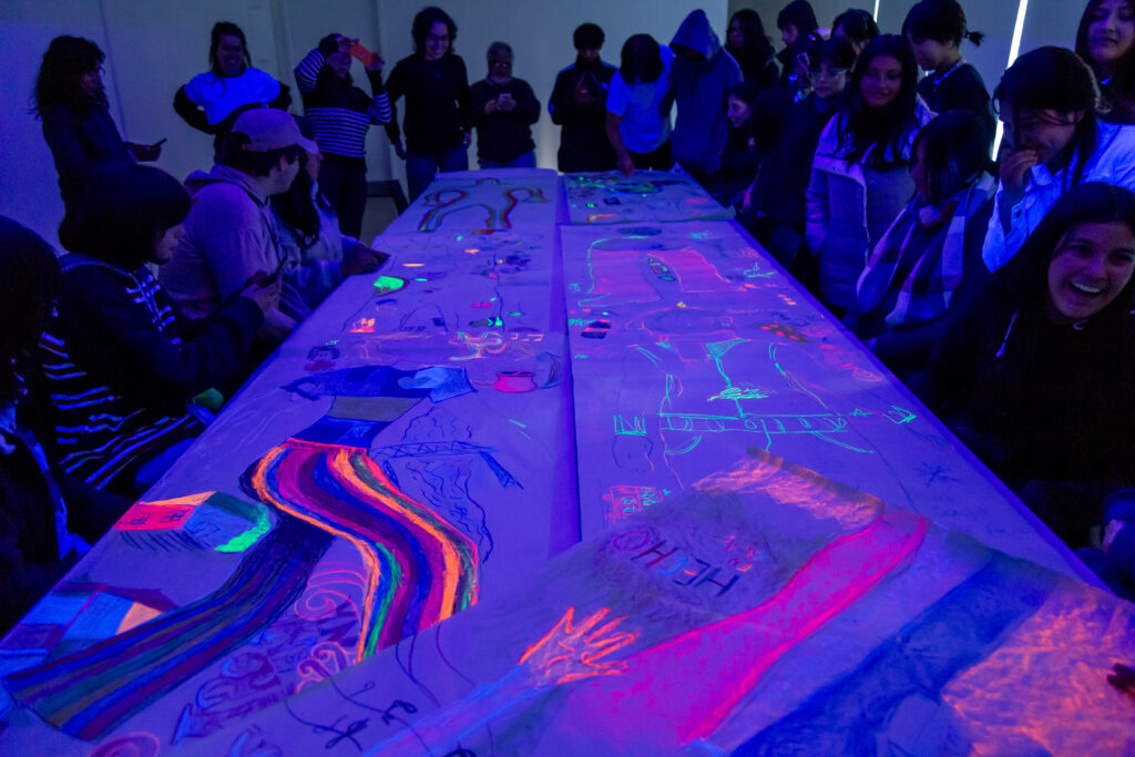 En la fotografía a color se observa un grupo de personas, en una sala ilumnada con una luz especial que destaca sus crayones fluor y la obra que realizaron con ellos en el marco de uno de los talleres continuados del Museo Moderno