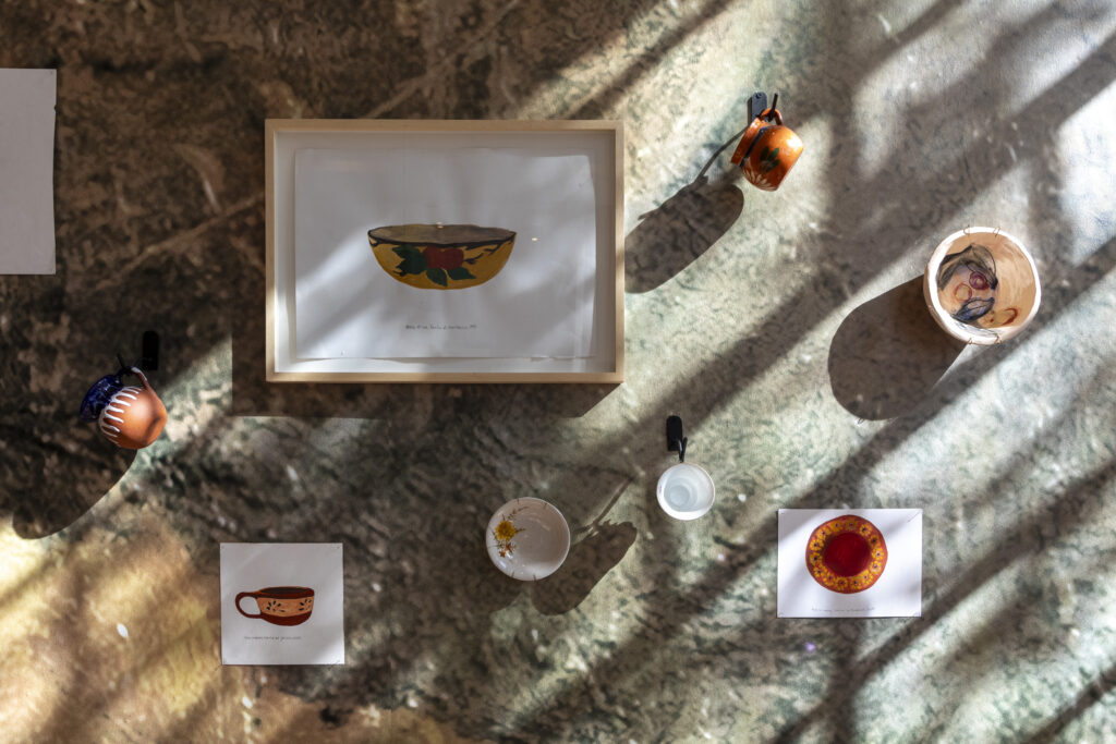 En la fotografía a color un cuadro, dibujos y tazas obras de Ana Gallardo en el marco de su exposición en el Museo Moderno "Materiales para la memoria"