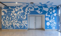 La imagen muestra el hall principal del Moderno, donde se encuentra la puerta de acceso a la sala Yapeyú, intervenido con un mural monocromático en color turquesa. Se trata de la exposición Alberto Passolini: Soñar a borbotones, en la cual, a partir de una gota, se despliega un mural que ocupa todas las paredes y el techo del lugar.