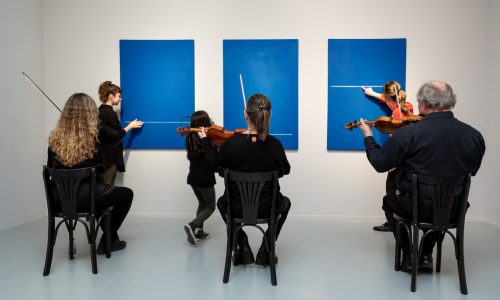 La fotografía muestra una activación en sala, en la que tres personas siguen con sus manos el recorrido de las líneas blancas que atraviesan tres cuadros de color azul. Detrás, tres músicos interpretan sonidos con sus violines.