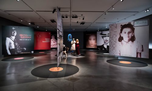 La imagen muestra la sala de la exposición Nacha Guevara: Aquí estoy donde se ven grandes paneles con fotografías de la artista, suspendidas arriba de unos discos de vinilo pegados en el suelo. En el centro de la imagen, se ven maniquíes con ropas, y de fondo, un video de gran formato donde se ve a Nacha cantando.