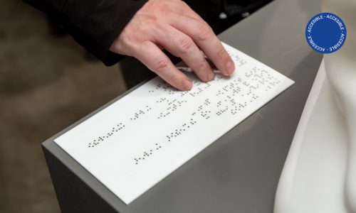 Foto muestra una vista accesible por la sala de educación en la que se ve una mano recorriendo la cédula en Braille de una obra de patrimonio