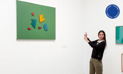 Fotografía a color muestra una sala de la exposición Moderno y MetaModerno donde se exhiben obras de la colección del museo y una persona usando Lengua de Señas.
