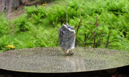 La fotografía muestra una piedra atada de un hilo, apoyada como péndulo sobre un disco plano agujereado, con un paisaje frondoso de fondo.