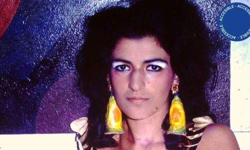 Fotografía a color: retrato en primer plano del rostro de la artista Dalila Puzzovio. La imagen muestra a una mujer joven, de cabello negro sobre los hombros y grandes ojos oscuros, luciendo unos aros muy voluminosos que contrastan con su tocado y con el fondo de la imagen donde predomina el azul brillante.