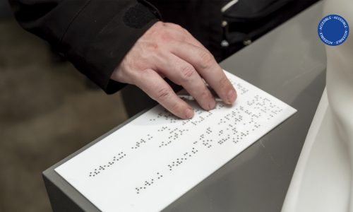 Foto muestra una vista accesible por la sala de educación en la que se ve una mano recorriendo la cédula en Braille de una obra de patrimonio