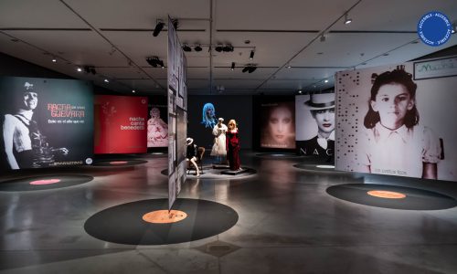 La imagen muestra la sala de la exposición Nacha Guevara: Aquí estoy donde se ven grandes paneles con fotografías de la artista, suspendidas arriba de unos discos de vinilo pegados en el suelo. En el centro de la imagen, se ven maniquíes con ropas, y de fondo, un video de gran formato donde se ve a Nacha cantando.