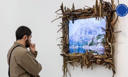 La foto muestra a una personas de espaldas viendo una TV de pantalla plana donde se proyecta un paisaje. El marco de la TV está cubierto de pequeños tronquitos de madera. La pieza es parte de la instalación de Antonio Villa en la sala de exposición del segundo subsuelo del Moderno.