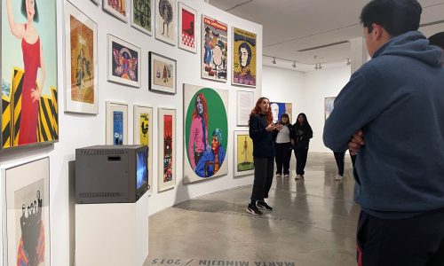 Fotografía tomada en la sala de exposición donde se ve la educadora del Museo frente a un grupo de visitantes. De fondo, hay pinturas coloridas de diferentes tamaños sobre una pared curva.