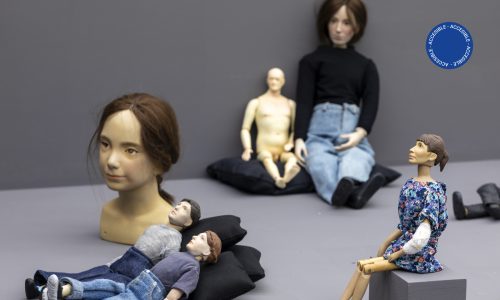 La imagen muestra muñecas de porcelana de diferentes tamaños; algunos de estos personajes tienen ropas y otros no. Están sentados o recostados sobre una base gris o sobre pequeños almohadones negros.