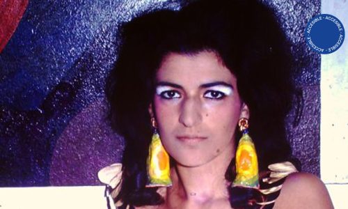 Fotografía a color: retrato en primer plano del rostro de la artista Dalila Puzzovio. La imagen muestra a una mujer joven, de cabello negro sobre los hombros y grandes ojos oscuros, luciendo unos aros muy voluminosos que contrastan con su tocado y con el fondo de la imagen donde predomina el azul brillante.